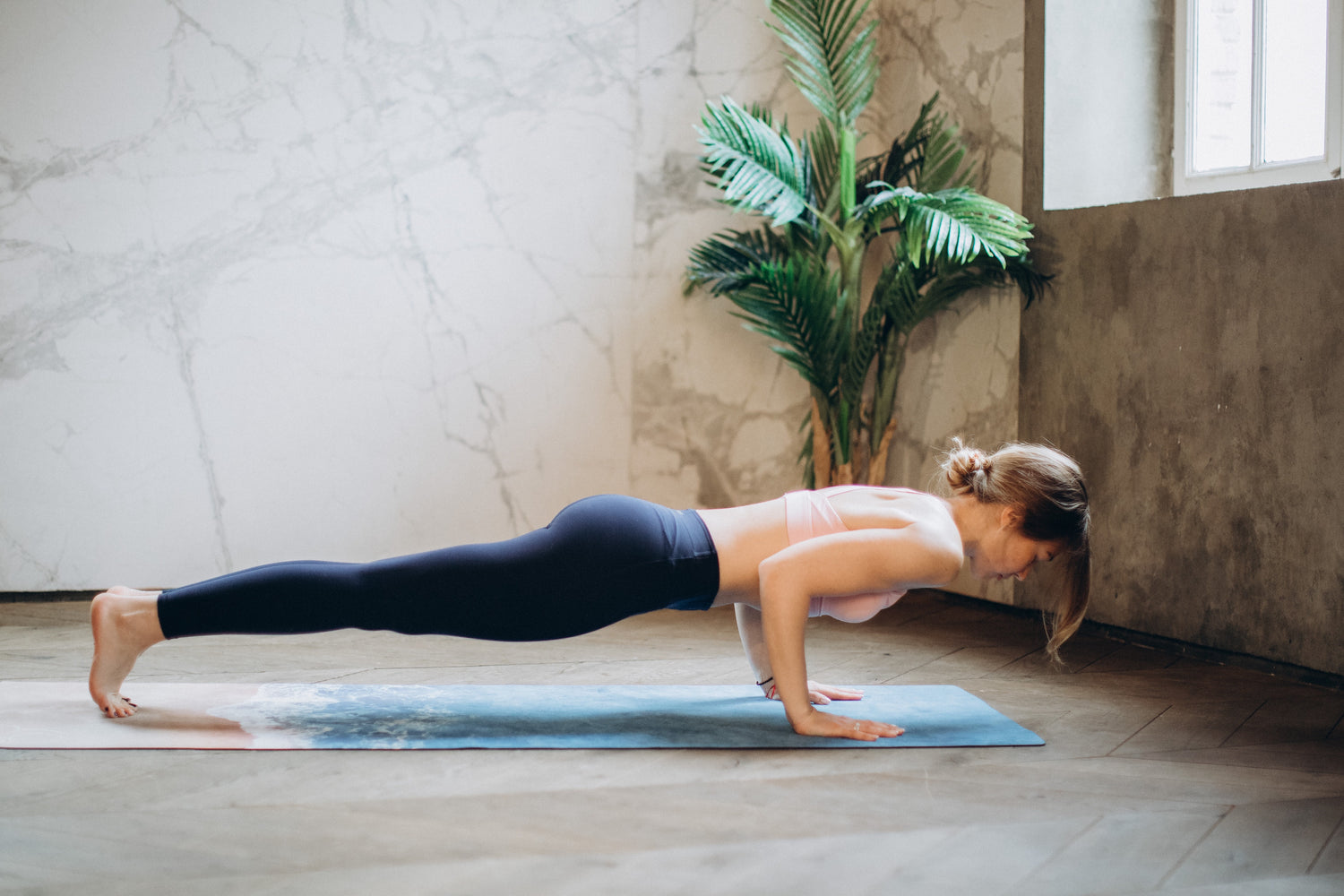girl in chaturanga yoga pose on yoga mat in neutral room with palm plant in corner
